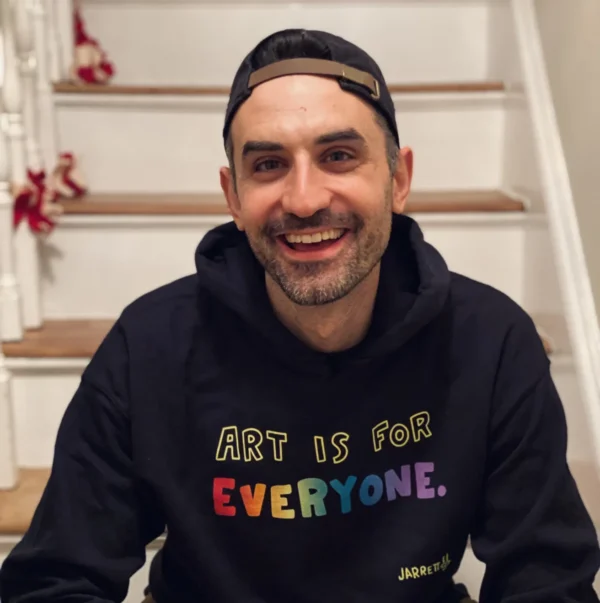 A man sits on wooden stairs, smiling, wearing a black hoodie that says 'Art is for everyone' in rainbow letters and a backwards cap.