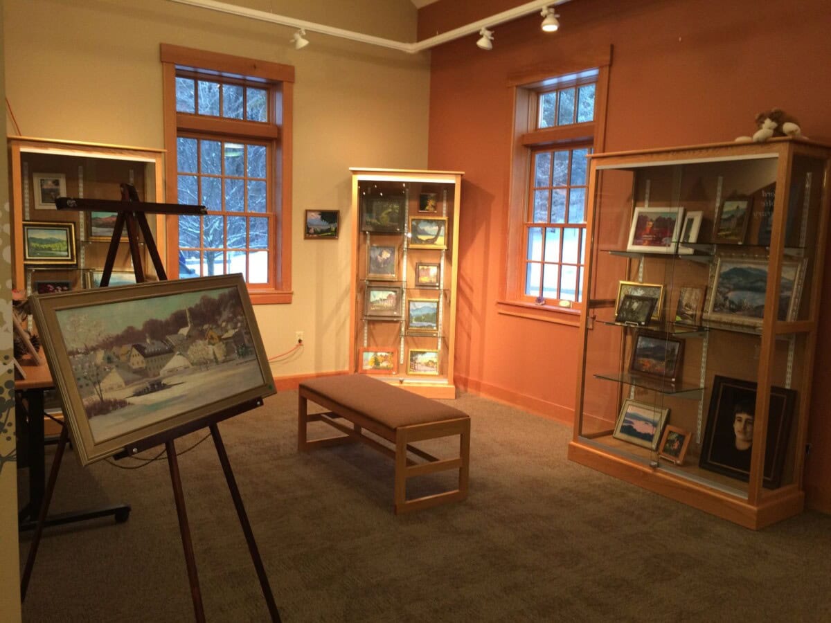 Art gallery room with paintings displayed on easels and inside glass cabinets, featuring two large windows and a wooden bench in the center.