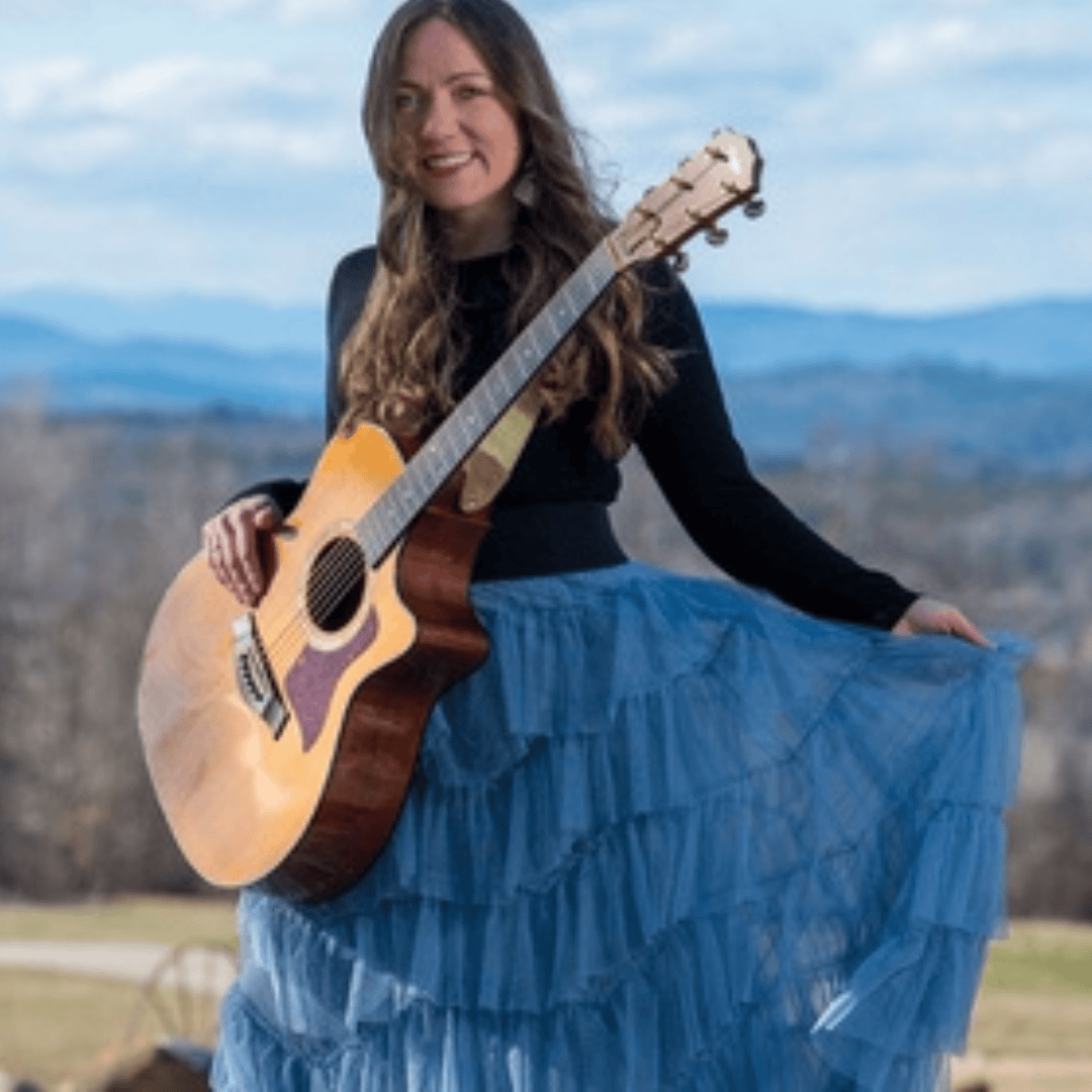 woman standing holding guitar in front of mountain landscape.