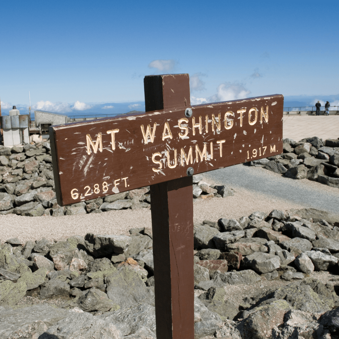 a wooden sign at the summit of mt. washington reads "mt. washington summit, 6,288 ft, 1,917 m" with rocky terrain and a viewing area in the background.
